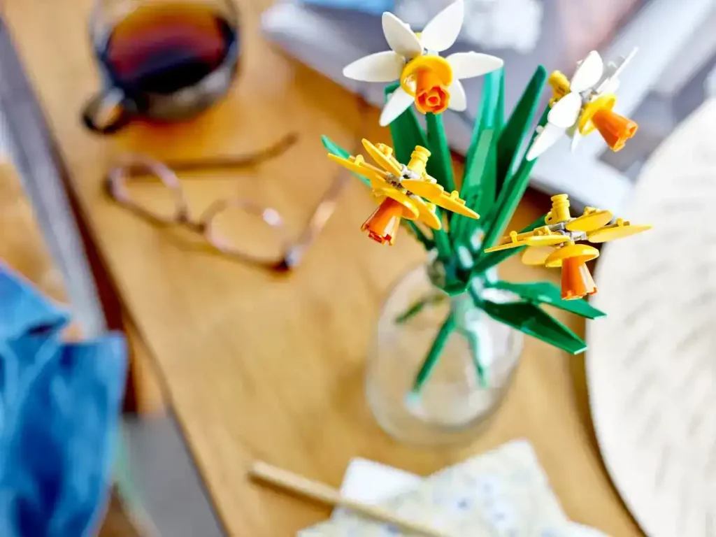 Close-up of LEGO Daffodils 40747 arranged in a glass vase on a wooden table next to a cup of tea and reading glasses