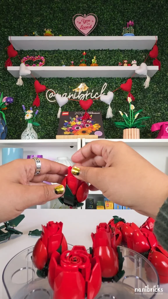 Hands holding a LEGO rose above a clear heart-shaped vase while planning a decorative flower arrangement.