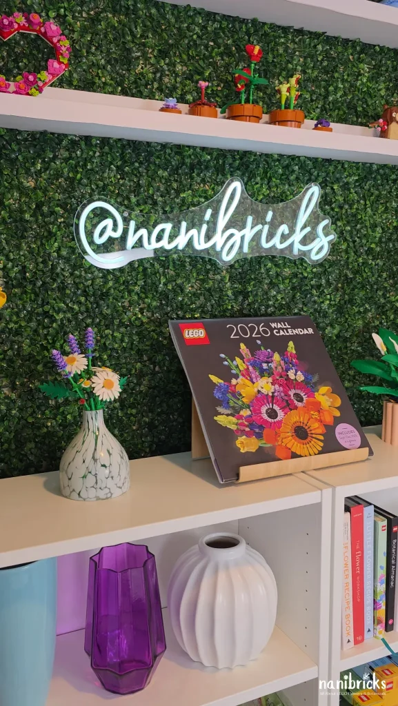 LEGO Botanicals Daisies 11508 displayed in a white vase on a shelf with LEGO 2026 wall calendar and greenery backdrop