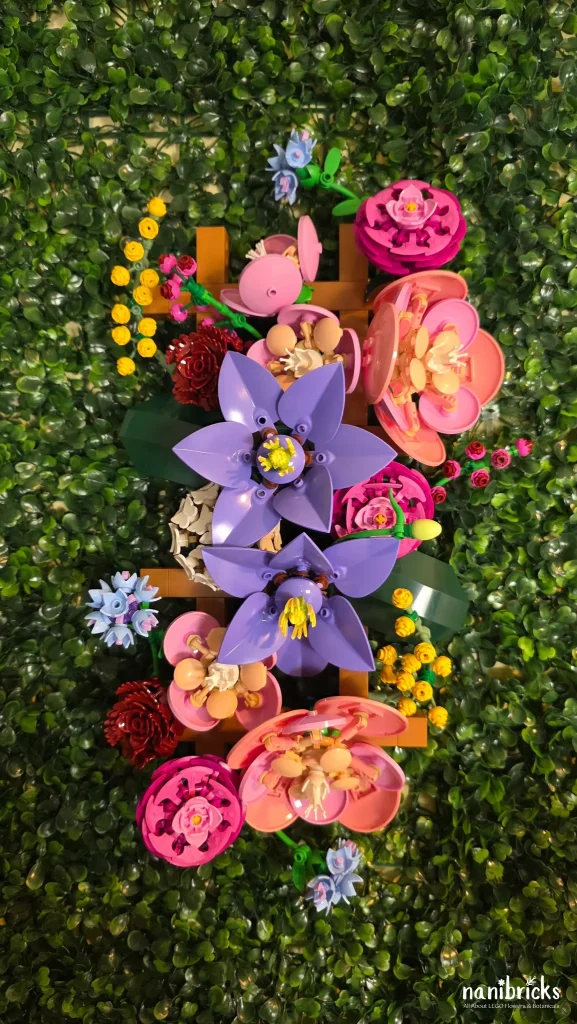 Vertical LEGO Botanicals Flower Wall (11503) mounted on greenery, showing camellias, clematis, ranunculus, roses, cornflowers, and mimosa arranged as wall art