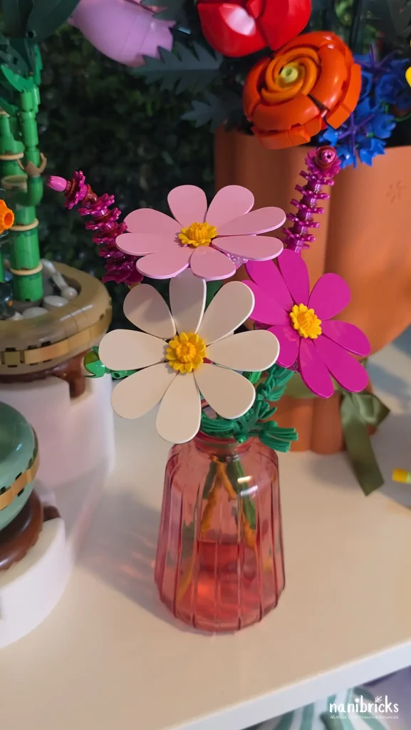 LEGO Botanicals Cosmos Flowers (11514) displayed in a pink glass vase on a shelf among other LEGO Botanicals arrangements.